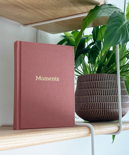 Hochwertiges Momente Erinnerungsbuch  mit Leineneinband und Goldprägung in Regal mit Pflanze, rosa